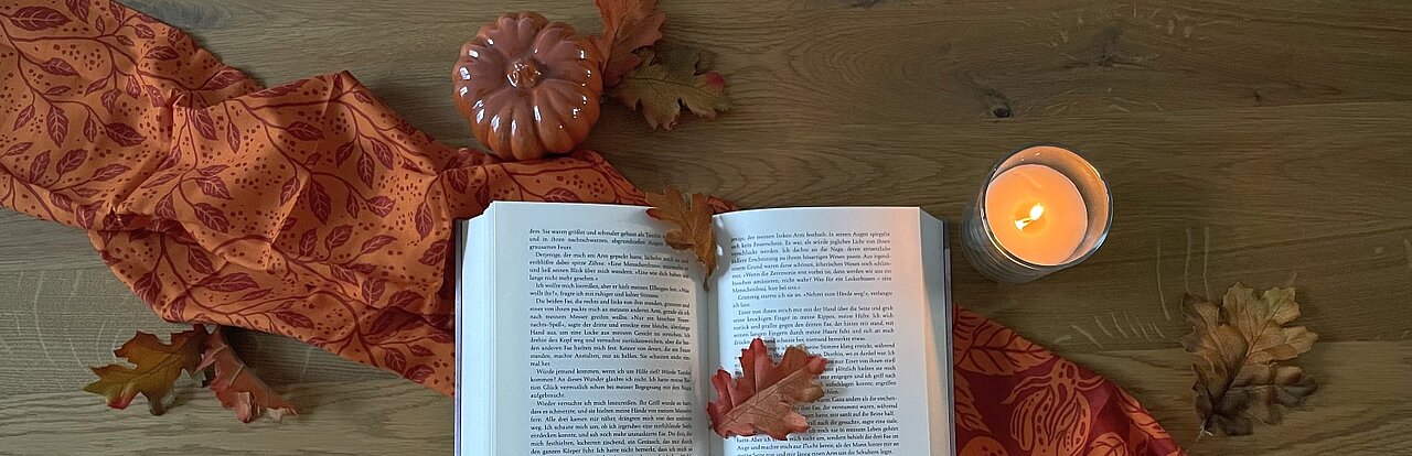 Auf diesem Bild sieht man ein aufgeschlagenes Buch umgeben von Herbst Dekor, darunter eine brennende Kerze, einen Kürbis und rote Laubblätter. 