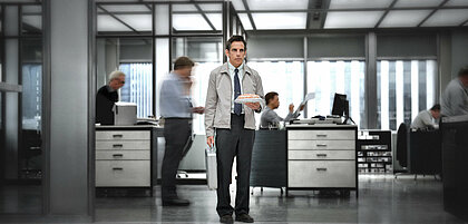 Zu sehen ist Walter Mitty, die Hauptfigur des Films, in einer typischen Büroszene. Er steht ruhig in der Mitte des Raums, während um ihn herum andere Personen in Bewegung sind.