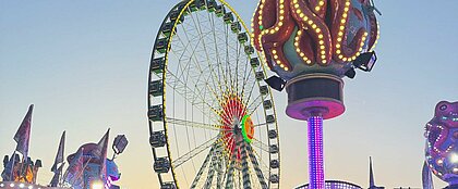 Auf dem Bild sieht man die Rheinkirmes. Im Vordergrund sieht man das Riesenrad und mehrere bunte Lichter beim Sonneuntergang.