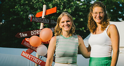 Auf dem Foto sind Isabell Nethke und Julia Riensche zu sehen. Sie stehen vor einem roten Schild und lächeln in die Kamera.