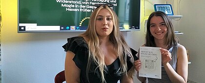 Auf dem Foto sind Nora und Blanca zu sehen. Sie sitzen nebeneinander und lächeln in die Kamera. Nora hält ein Flyer zu der Ausstellung hoch.
