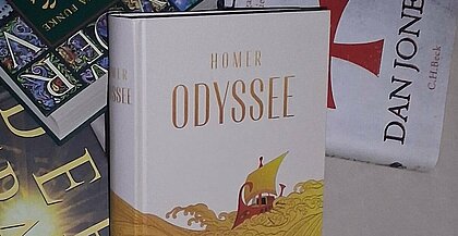 Auf dem Bild sieht man verschiedene Bücher auf einem Haufen liegen. Zentral liegt eine Ausgabe von Homers Odyssee.