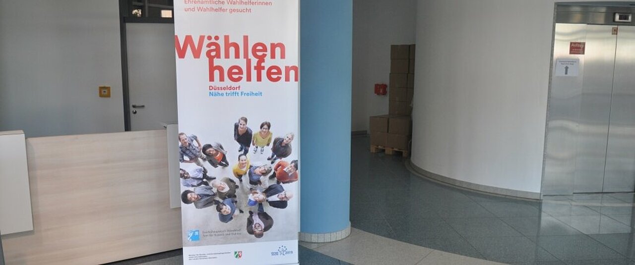 Es ist ein Foto vorhanden. Zu sehen ist ein Roll-Up auf dem in roter Schrift “Wählen helfen” steht.