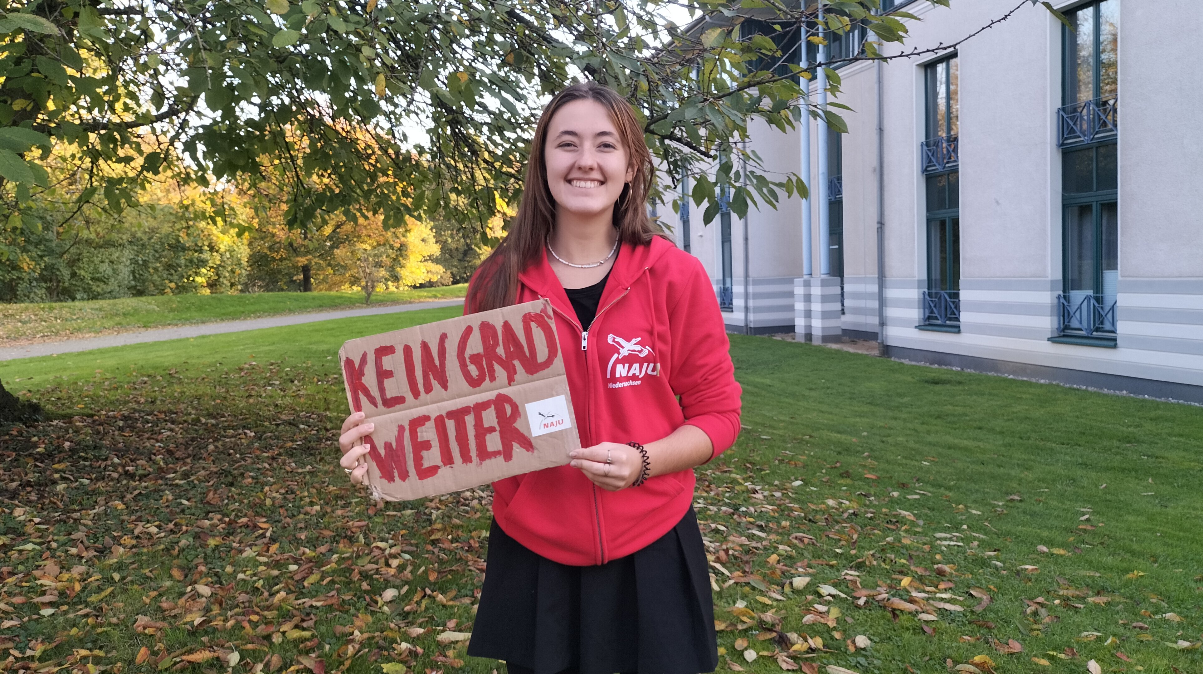 Auf dem Foto ist Yamina Probst zu sehen. Sie steht draußen auf einer Wiese und hält ein Schild mit der Aufschrift "Kein Grad weiter" hoch.