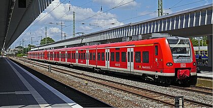 Zu sehen ist ein roter Zug, der auf Gleisen vor einem Bahnsteig steht. 