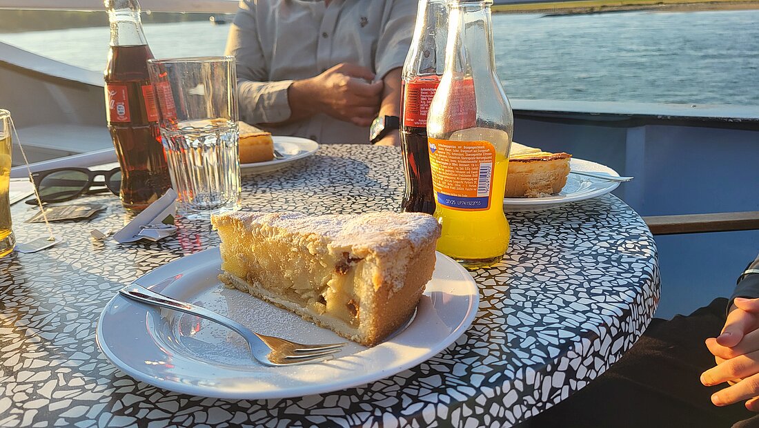 Auf dem Bild sieht man ein Stück Apfelkuchen auf dem Tisch am Sonnendeck des Schiffs.