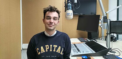 Auf dem Foto ist Josua Wienen zu sehen. Er steht im Studio, neben einem Mikrofon und lächelt in die Kamera.