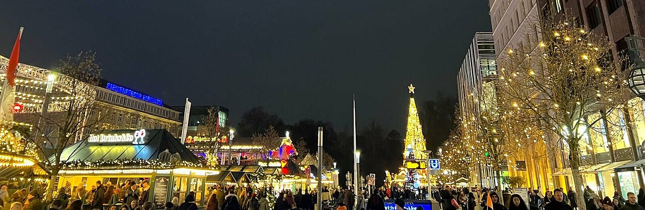 Auf dem Bild ist ein Weihnachtsmarkt in Düsseldorf zu sehen. Auf dem Bild sind viele Menschen und im Hintergrund ist ein großer Weihnachtsbaum zu erkennen.