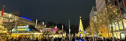 Auf dem Bild ist ein Weihnachtsmarkt in Düsseldorf zu sehen. Auf dem Bild sind viele Menschen und im Hintergrund ist ein großer Weihnachtsbaum zu erkennen.