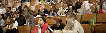 Auf dem Fotos sind Studierende in einem Hoersaal zu sehen. Sie blicken nach vorne.