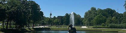 Park mit Fontäne auf einem Gewässer, im Hintergrund der Fernsehturm