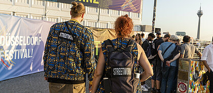 Man sieht zwei Streetworker:innen, die an der Rheinuferpromenade in der Düsseldorfer Altstadt unterwegs sind.