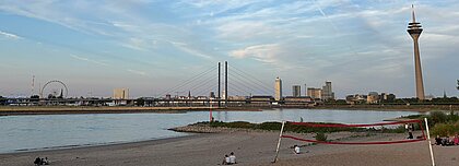 Auf dem Foto sieht man die Düsseldorfer Skyline vom Paradiesstrand aus. Der Himmel ist blau und auf dem Sand sitzen ein paar Menschen.