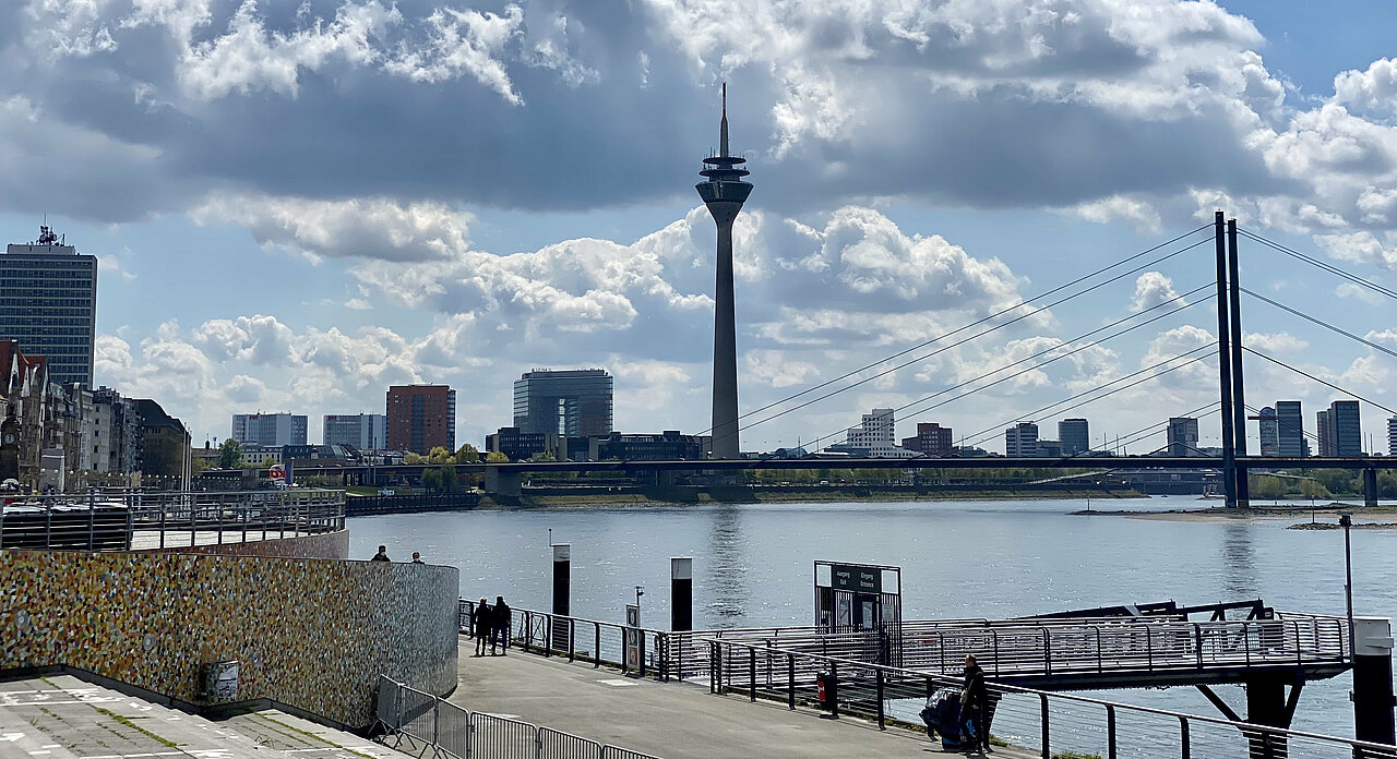 Auf dem Foto ist die Skyline von Düsseldorf mit Rheinturm zu sehen. Im Vordergrund ist der Rhein zu sehen. 