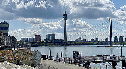 Auf dem Foto ist die Skyline von Düsseldorf mit Rheinturm zu sehen. Im Vordergrund ist der Rhein zu sehen. 