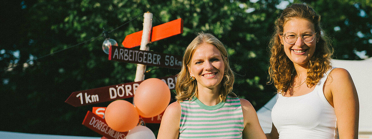 Auf dem Foto sind Isabell Nethke und Julia Riensche zu sehen. Sie stehen vor einem roten Schild und lächeln in die Kamera.