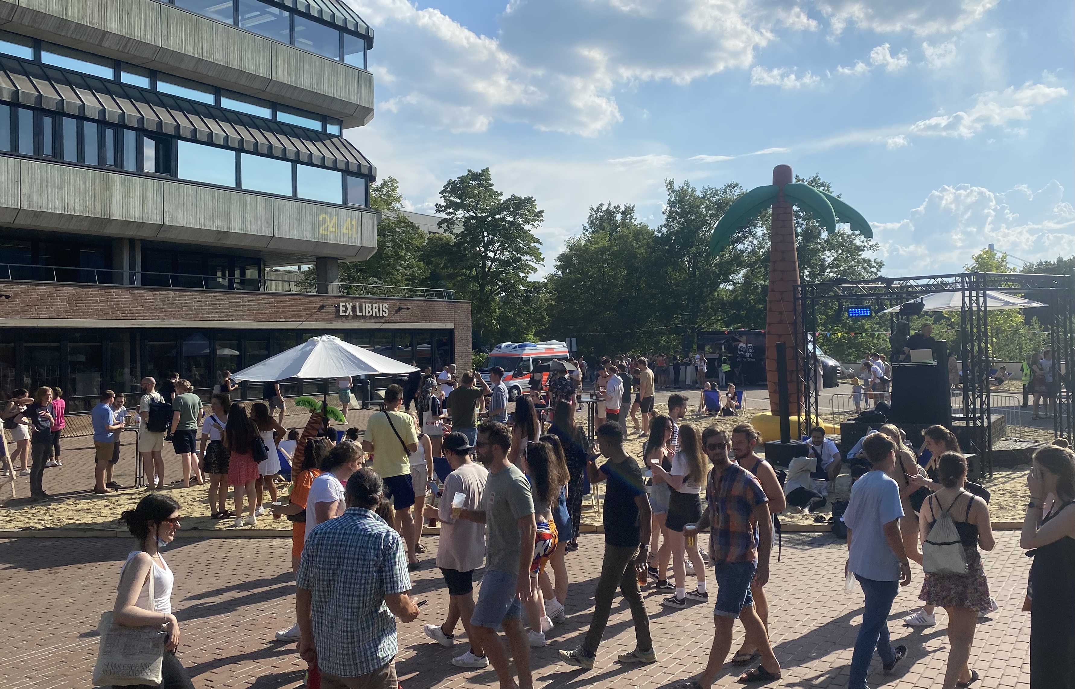 Man sieht mitten auf dem Campus der Heinrich-Heine-Universität eine Sandflächen mit Schirm, einer kleinen Luftpalme darunter auf der linken Seite und rechts eine große Luftpalme und weiter rechts eine kleine Bühne für den DJ. Auf der Sandfläche, dahinter und davor ist eine Menschenmenge versammelt. 