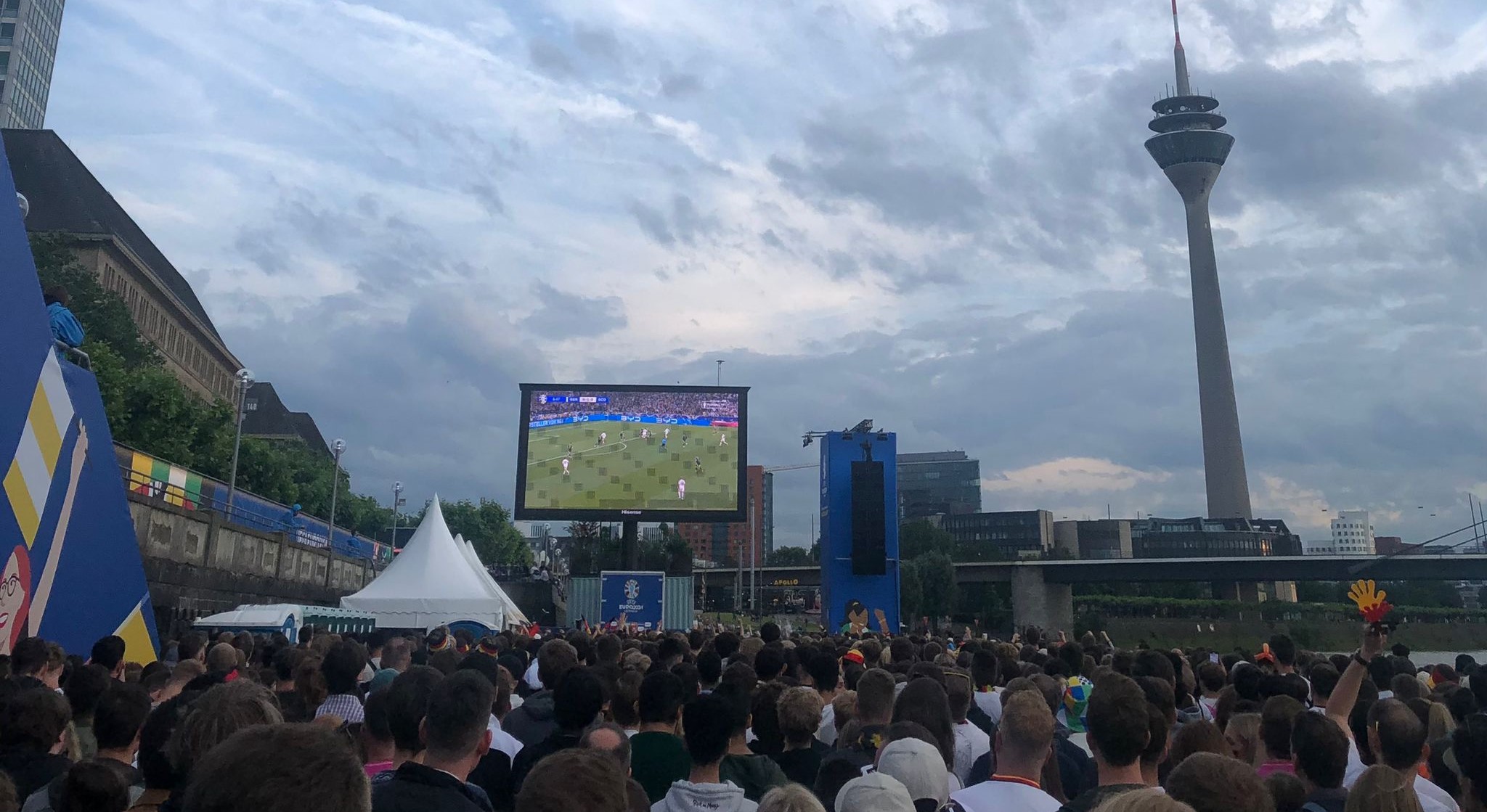 Auf dem Bild ist die Fan Zone am Rheinufer in Düsseldorf zu sehen. Auf einem großen Bildschirm läuft ein Deutschland Spiel. Es sind viele Leute zusehen, die gekommen sind um das Spiel anzusehen.